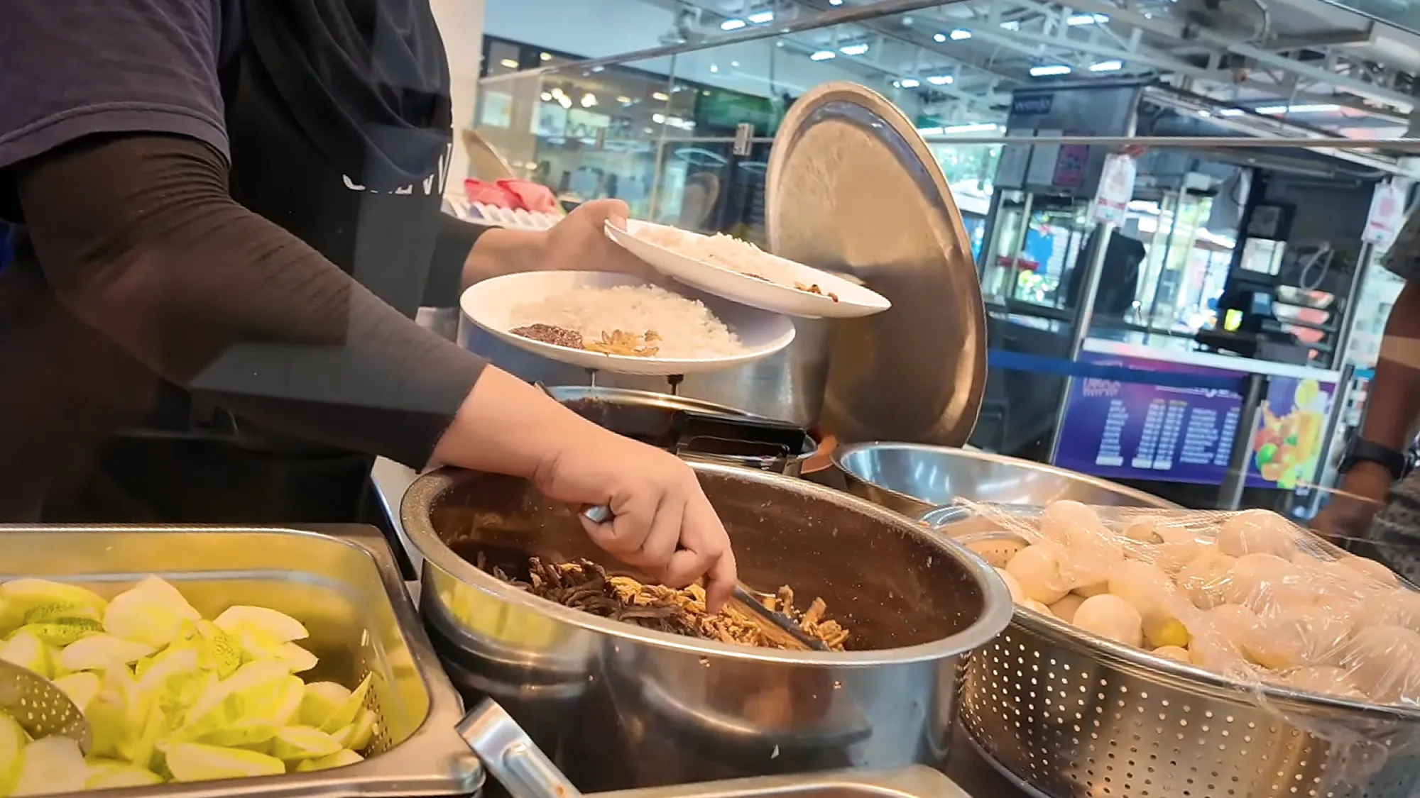 Staff at Wanjo's counter building a plate of nasi lemak, adding anchovies beside trays of cucumber and eggs