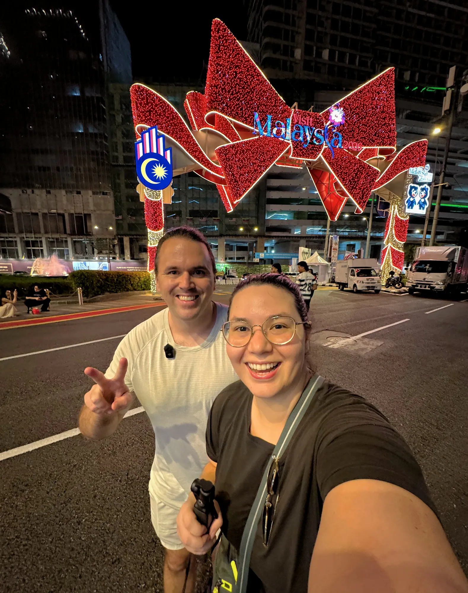 Bernie and Cream taking a night selfie under the lit-up red Visit Malaysia 2026 arch in Bukit Bintang, Kuala Lumpur.