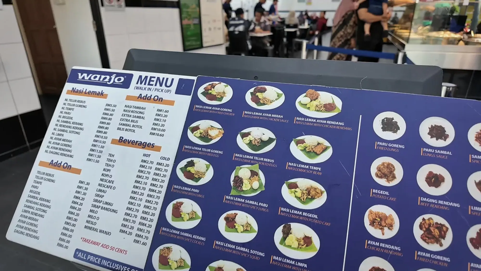 The Wanjo menu board listing nasi lemak plates, add-ons, and drinks with prices in ringgit, with the dining room behind it.