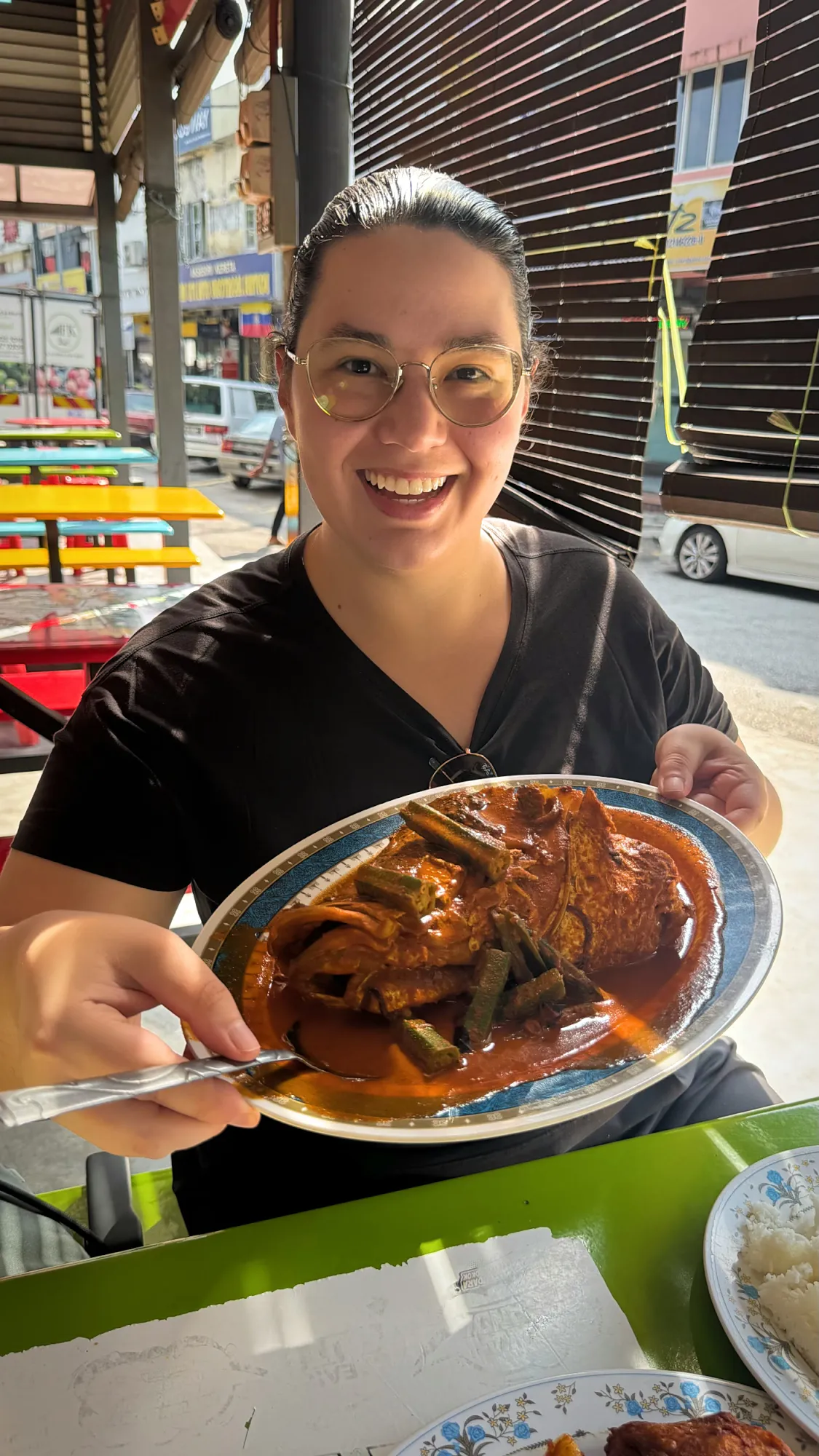 Bernie smiling and holding up a platter of fish head curry with okra at an outdoor table at Anuar Curry Fish Head in Bangsar, Kuala Lumpur.