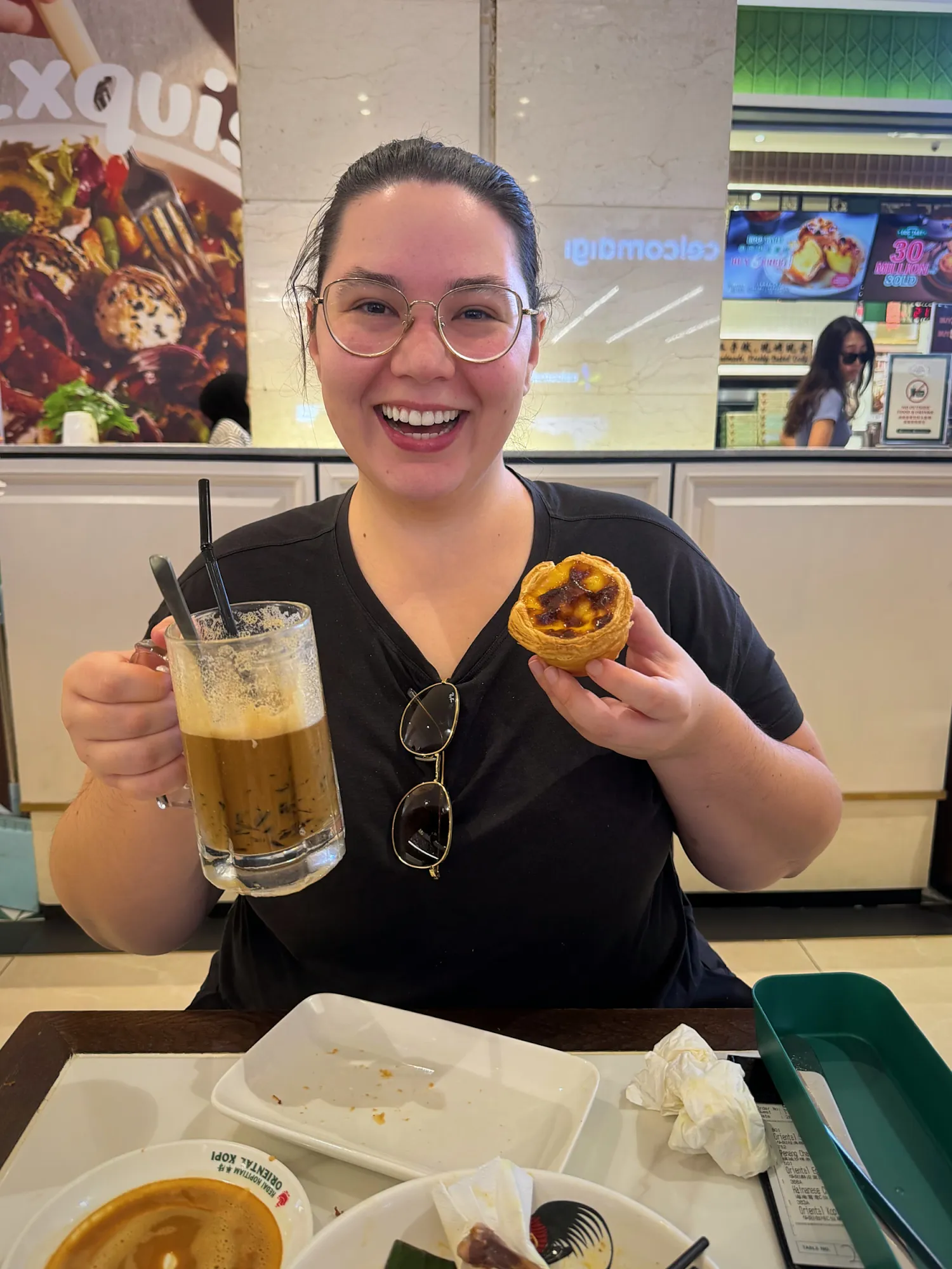 A smiling woman holding up a mug of iced kopi and an egg tart at Oriental Kopi in Kuala Lumpur.