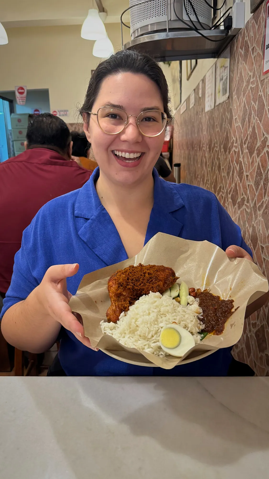 Holding up a Village Park nasi lemak with crispy fried chicken, sambal, cucumber, and a boiled egg on brown paper