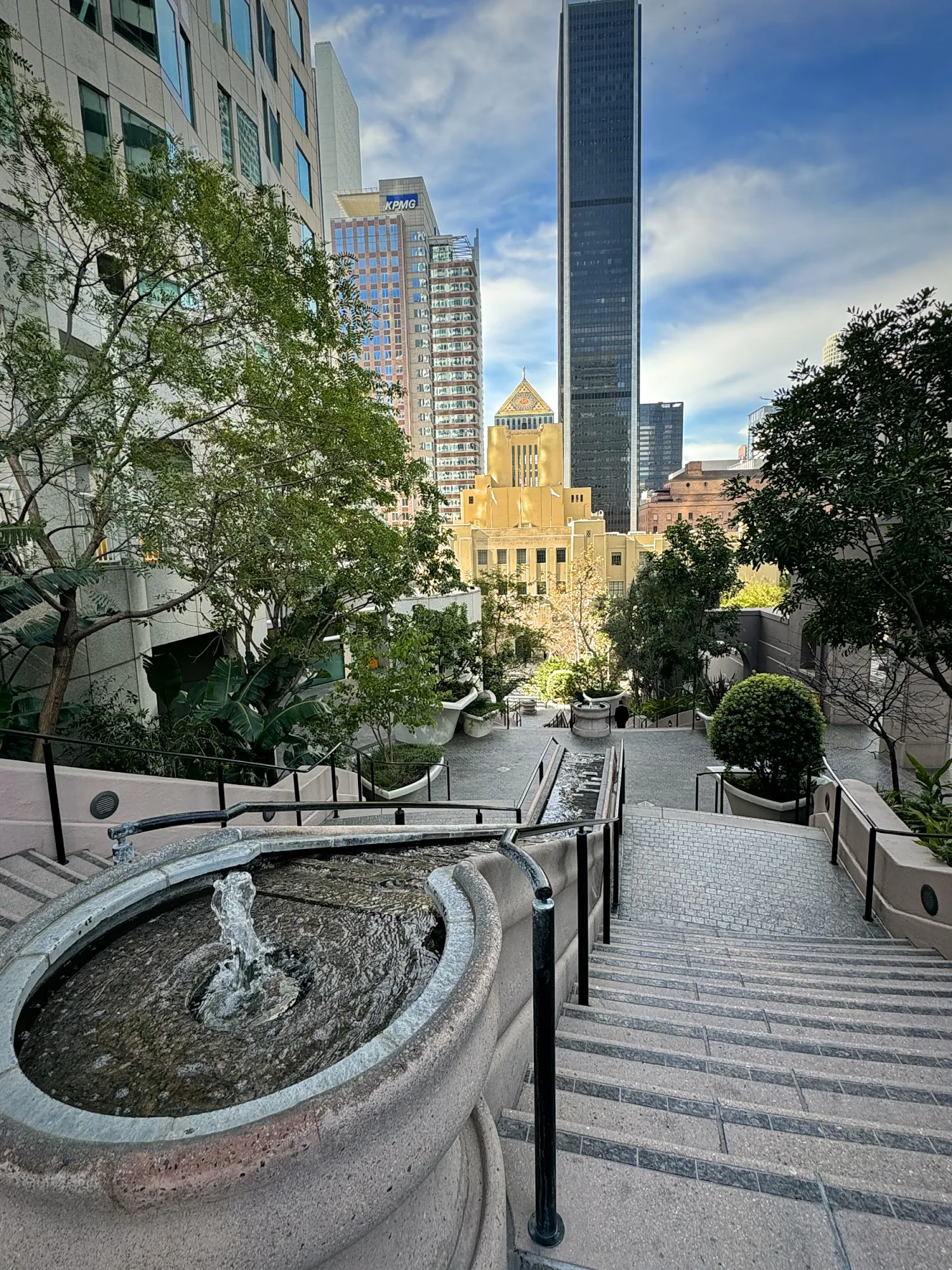 A fountain at the top of the Bunker Hill Steps in Downtown LA, with trees, office towers, and the tiled tower of the Central Library behind.