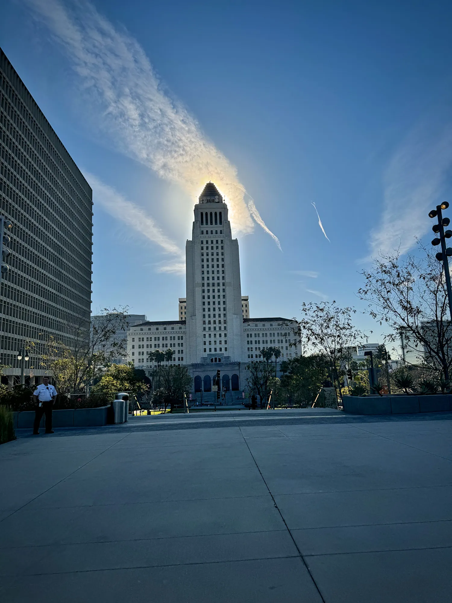 The white tower of Los Angeles City Hall at the end of a wide paved plaza in Downtown LA, under a bright blue sky.