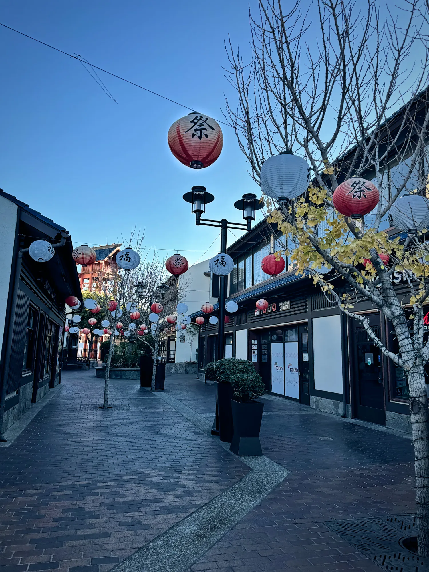 Red and white paper lanterns strung over the brick lane of Japanese Village Plaza in Little Tokyo, Los Angeles, with the red fire lookout tower behind them.
