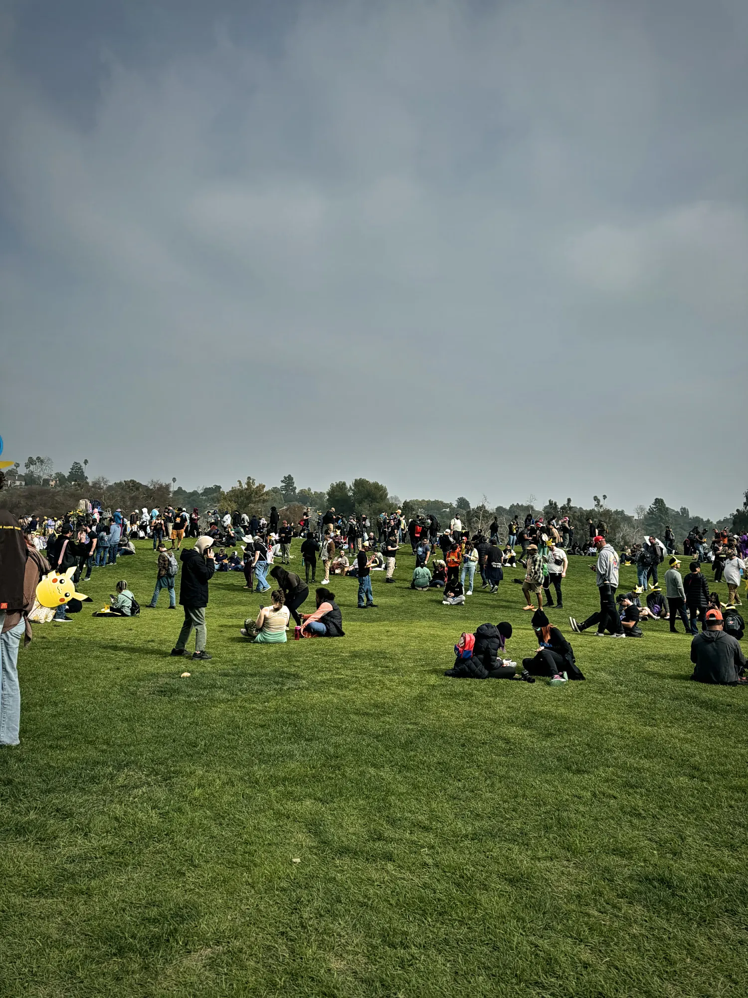 Pokémon GO players sitting and walking across a wide grassy hill under a grey sky at the Rose Bowl during GO Tour.