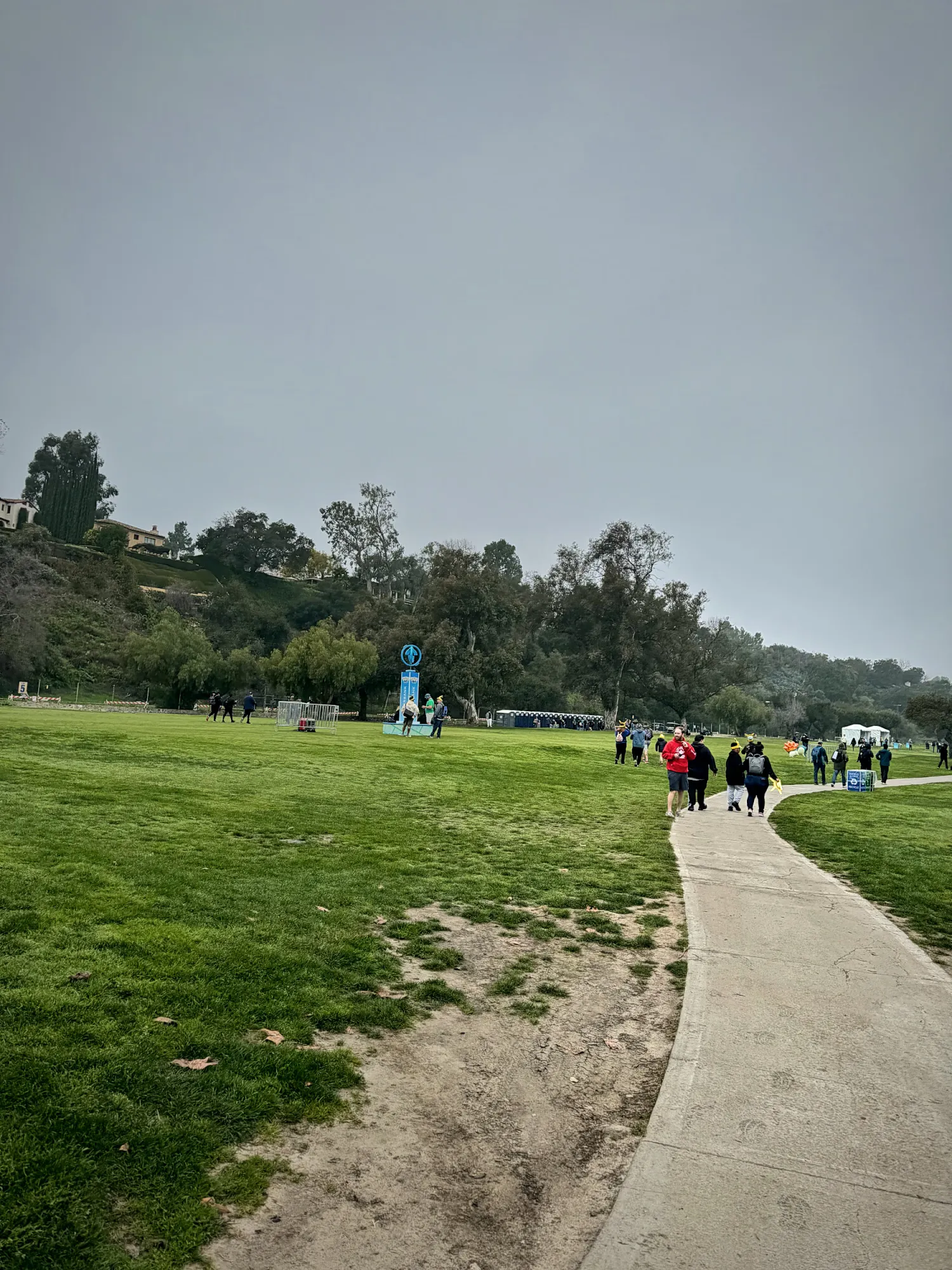 A paved path curving across the grass at the Rose Bowl in Pasadena on a gray morning, with Pokémon GO Tour trainers walking toward the event signs in the distance.