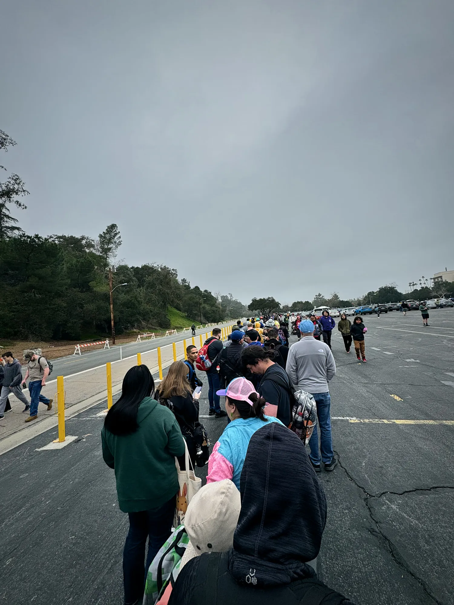 A long line of Pokémon GO Tour trainers in hoodies and caps waiting on a closed road beside a Rose Bowl parking lot on a gray morning in Pasadena.