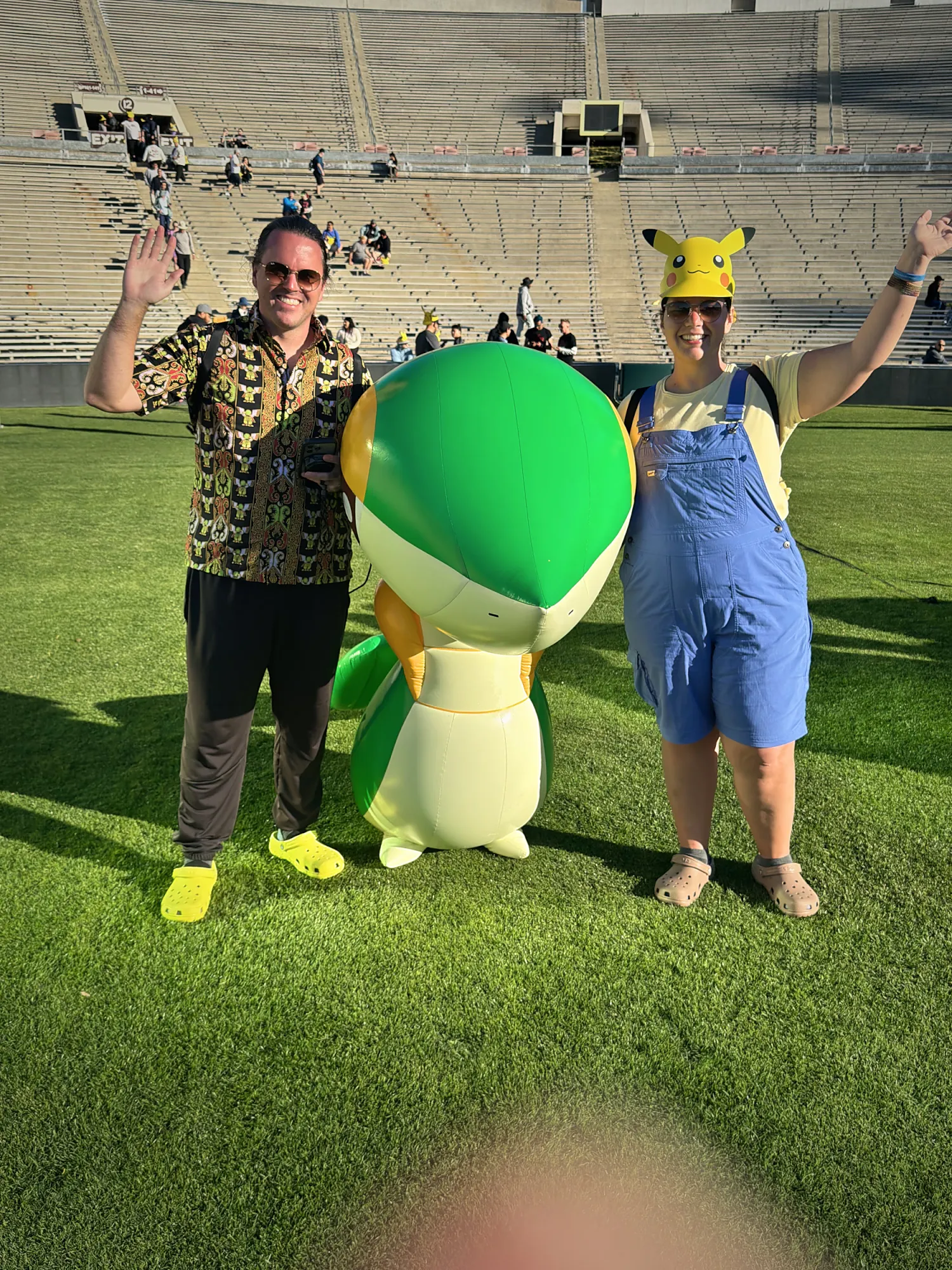 Bernie and Cream waving next to a giant Snivy on the field inside Rose Bowl Stadium at Pokémon GO Tour Los Angeles, with the stands behind them.