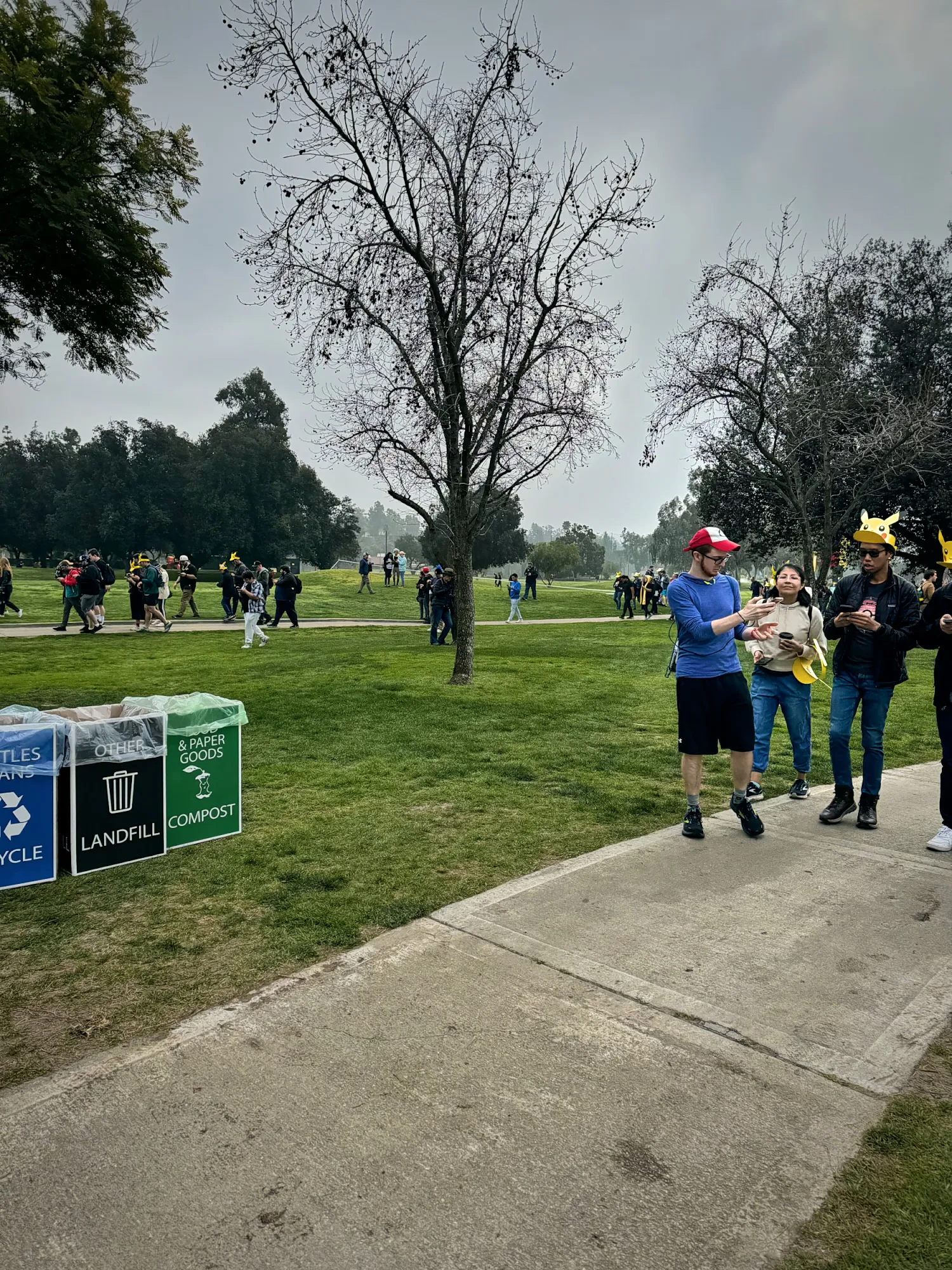 Trainers in Pikachu hats walking a paved path between lawns and bare trees at the Rose Bowl, next to trash, recycling, and compost bins.