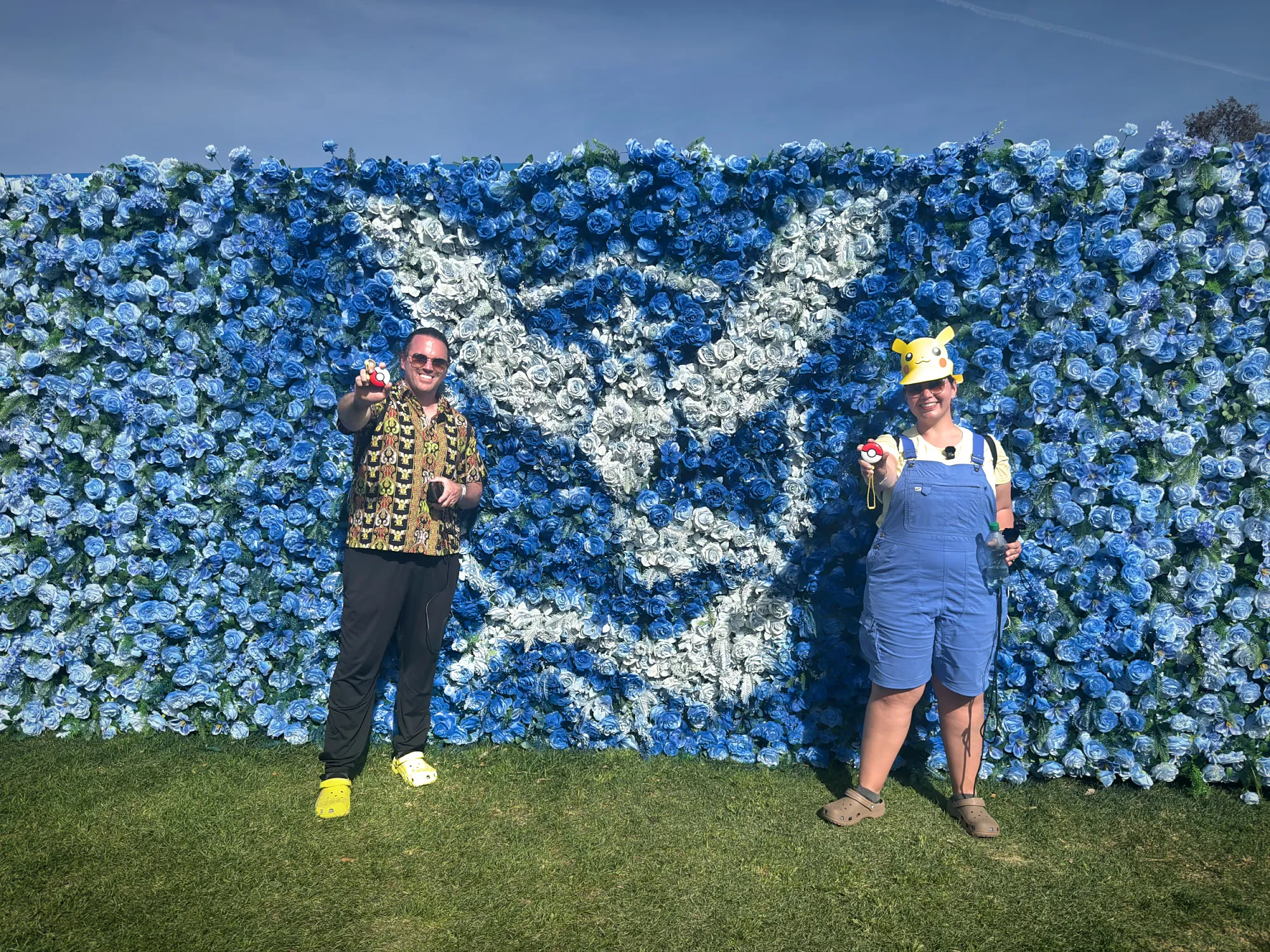 Bernie and Cream holding up Poké Balls in front of a blue Team Mystic flower wall at Pokémon GO Tour at the Rose Bowl in Pasadena.