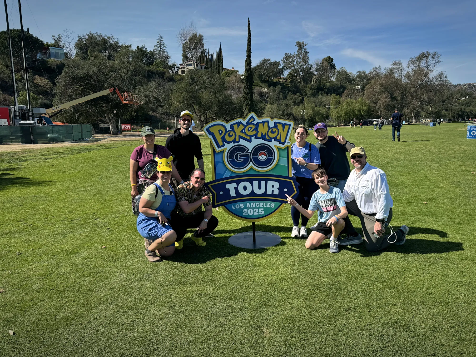 A group of Pokémon GO friends posing around a Pokémon GO Tour sign on the grass in Pasadena.