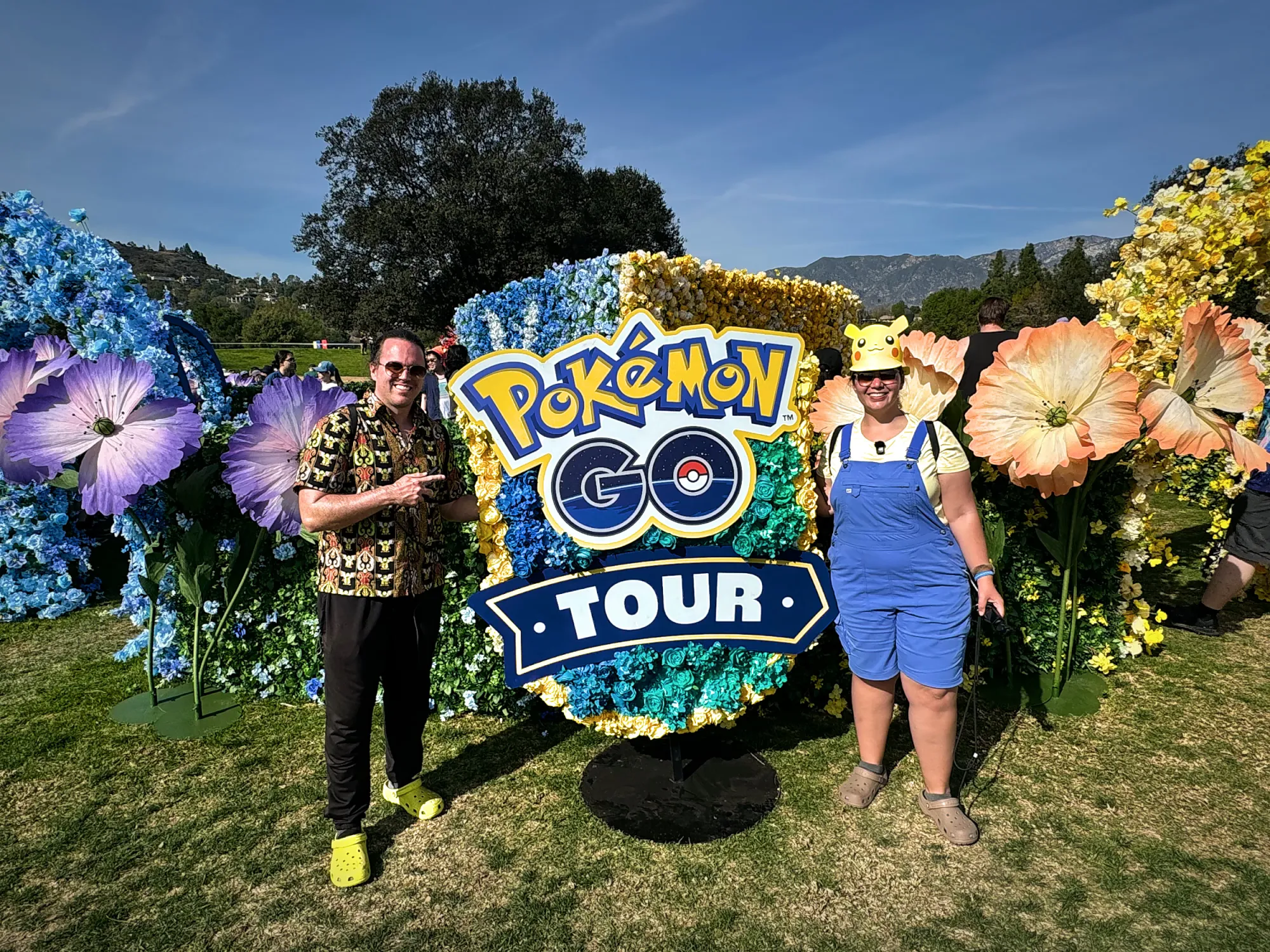 Bernie and Cream standing on either side of a Pokémon GO Tour sign surrounded by giant flowers at the Rose Bowl, one of them in a Pikachu visor.