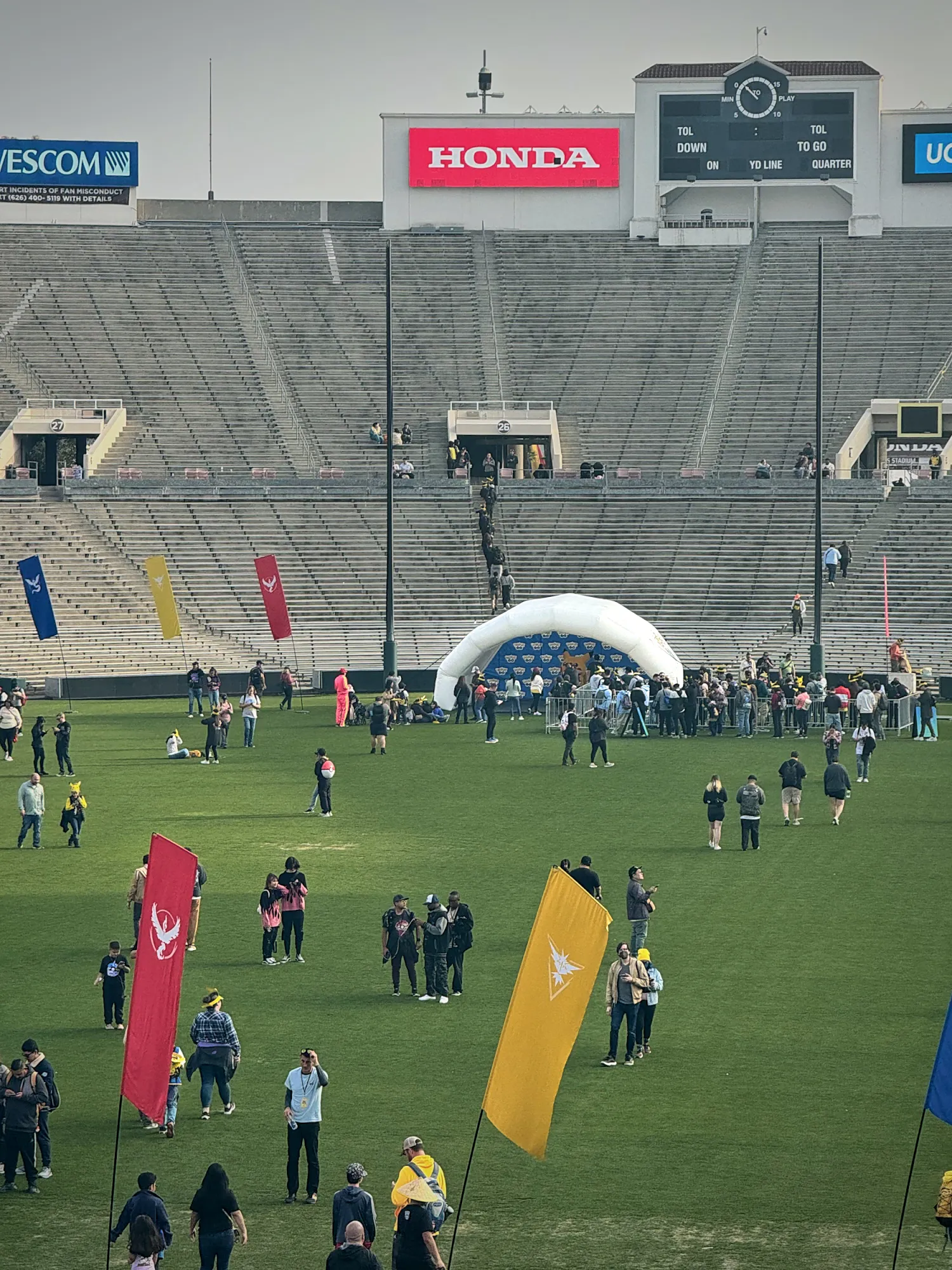 Trainers walking across the grass inside Rose Bowl Stadium past tall red, yellow, and blue Pokémon GO team flags, with a line at a white inflatable photo arch below the empty stands.