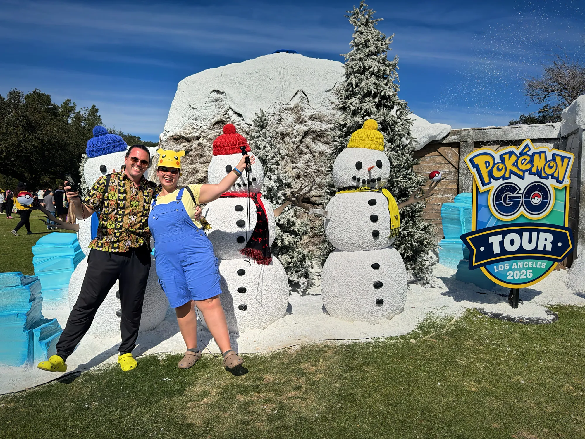 Bernie and Cream posing with snowmen in knit hats at the winter habitat of Pokémon GO Tour at the Rose Bowl, next to a GO Tour Los Angeles 2025 sign.