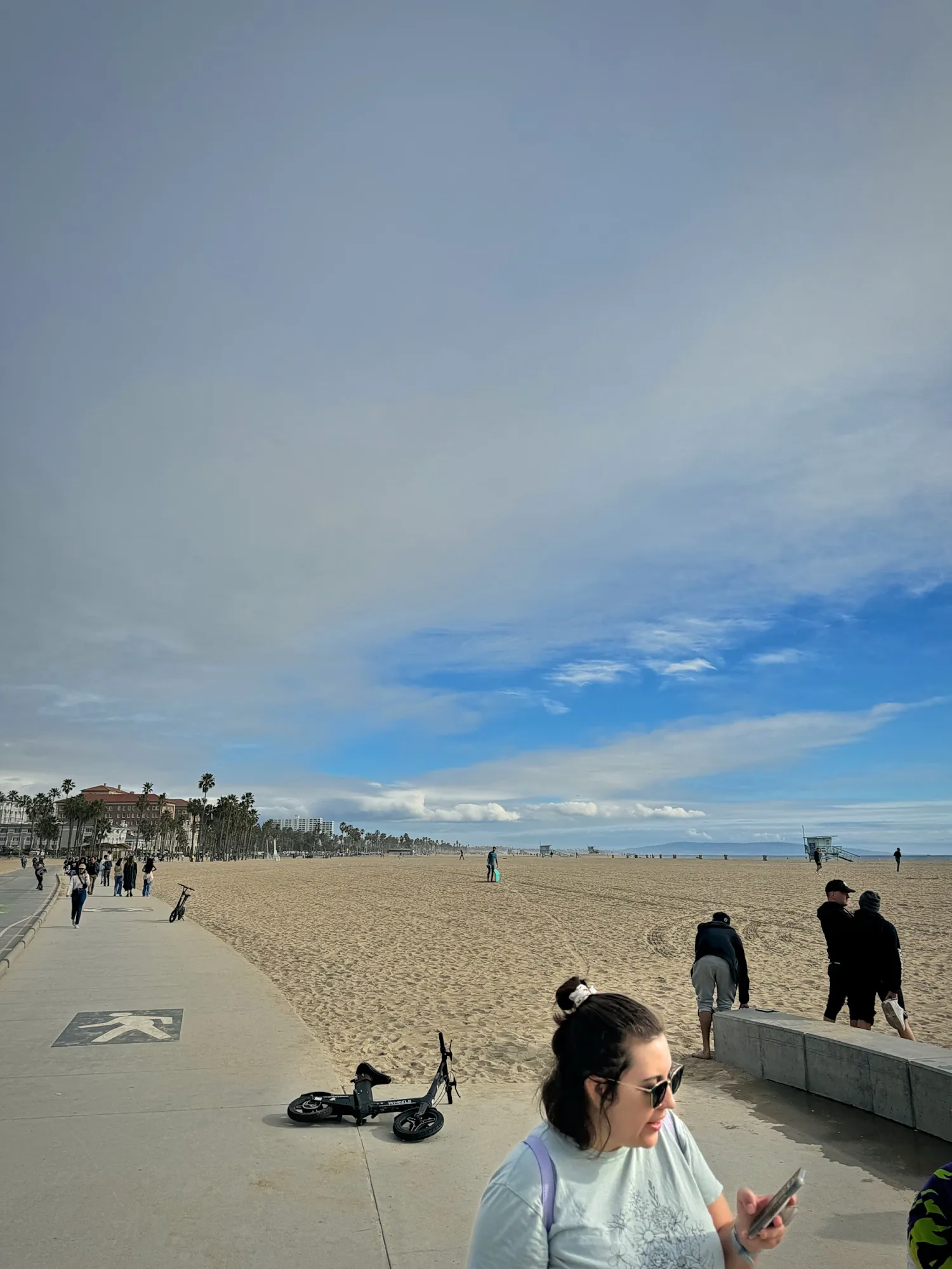 A woman in sunglasses checks her phone on the beach path in Santa Monica, with a wide sandy beach under a cloudy sky.
