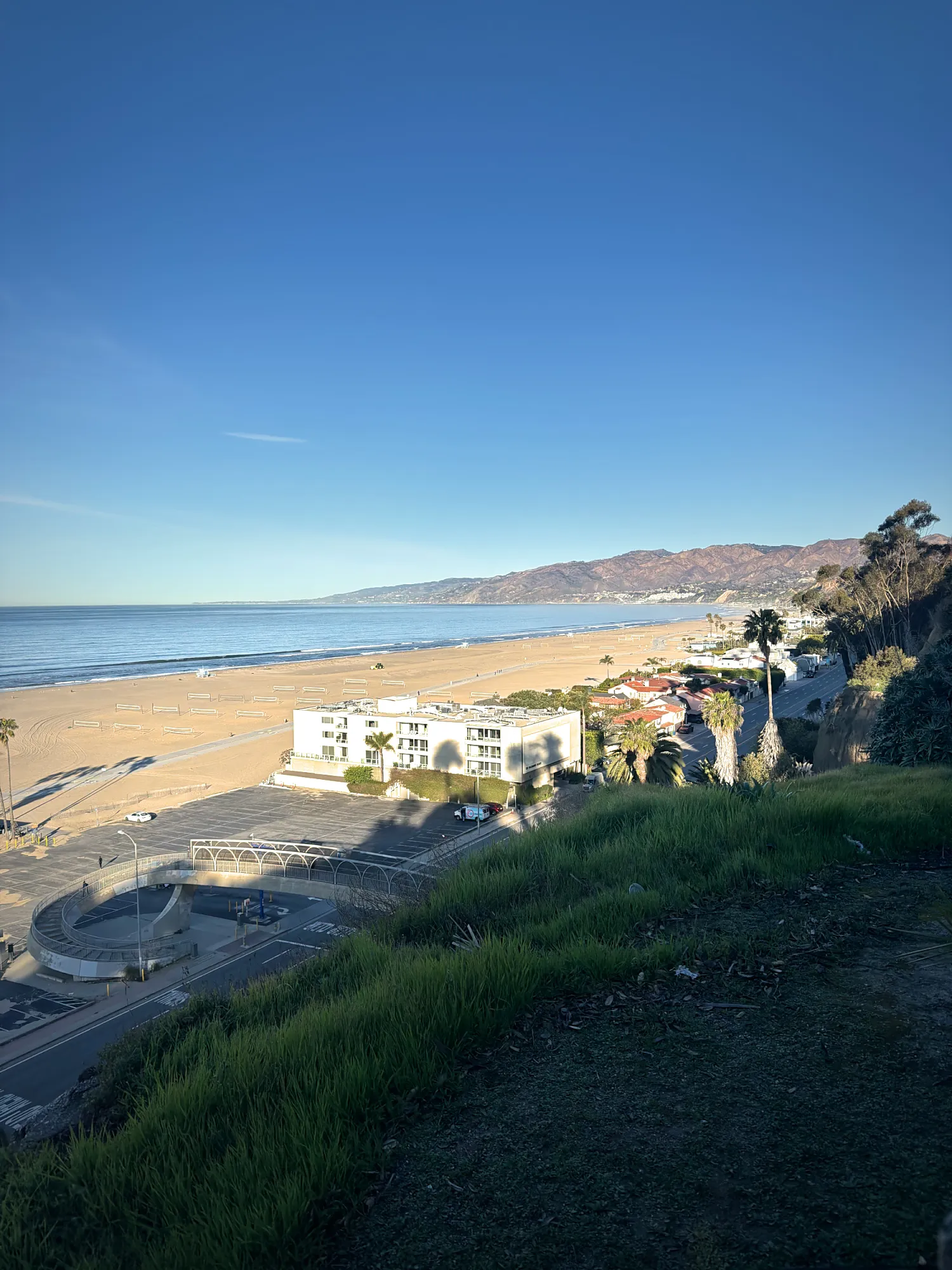 The wide sandy beach in Santa Monica and the mountains toward Malibu under a clear blue sky, seen from the grassy clifftop above the coast road.