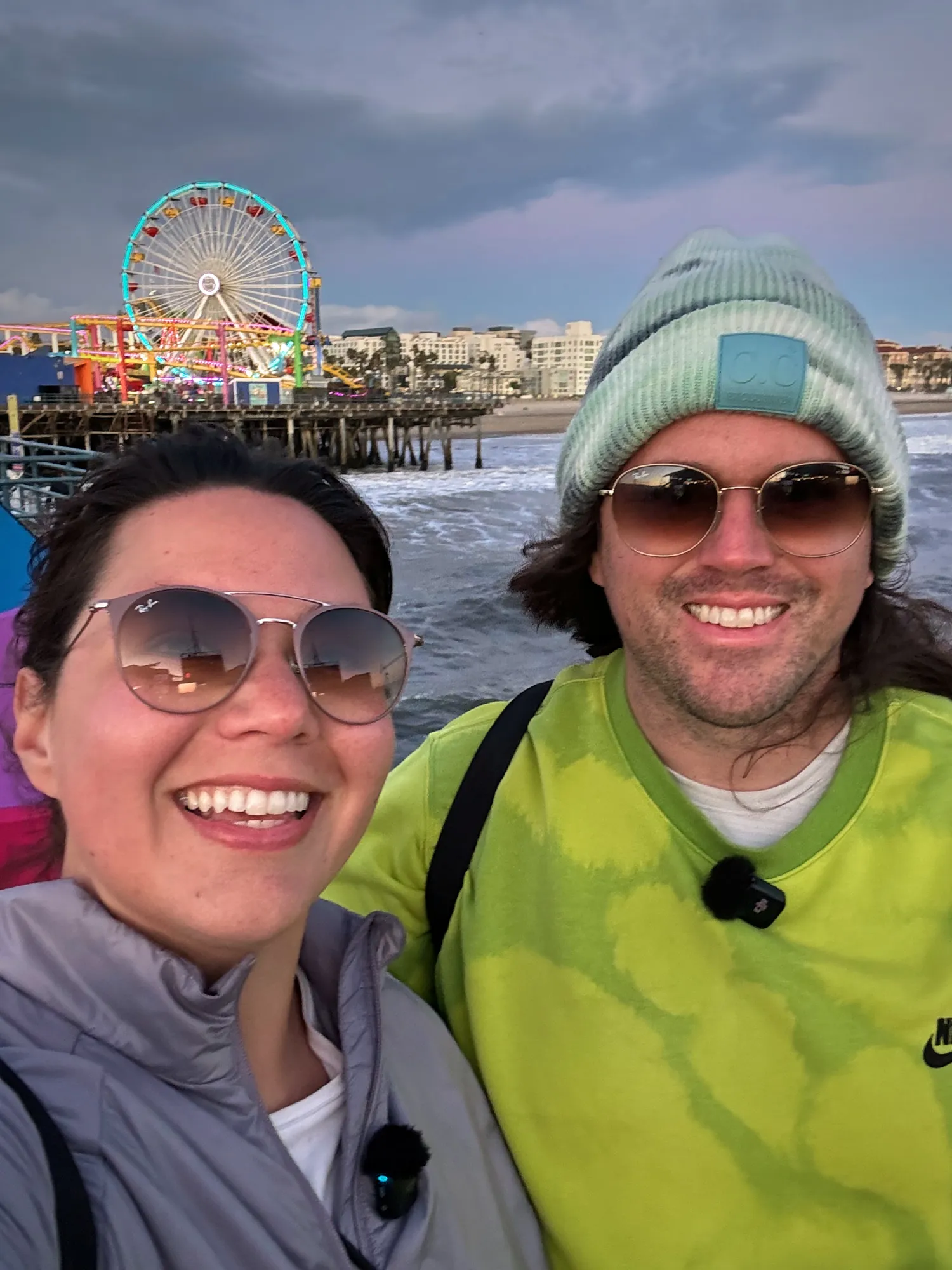 Bernie and Cream smiling for a selfie in sunglasses, with the Ferris wheel on Santa Monica Pier and the beach behind them on a cloudy day.