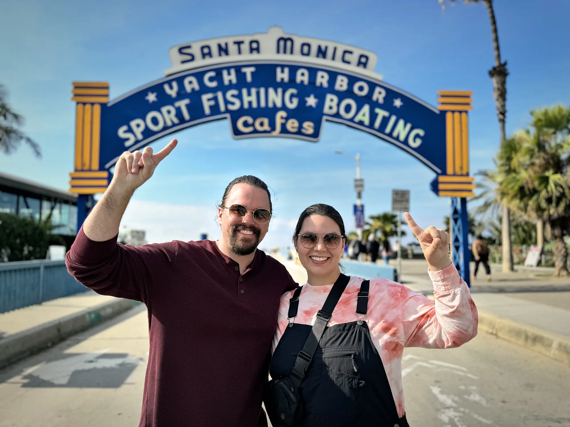 Bernie and Cream pointing up at the Santa Monica Yacht Harbor sign at the entrance to Santa Monica Pier on a sunny day.
