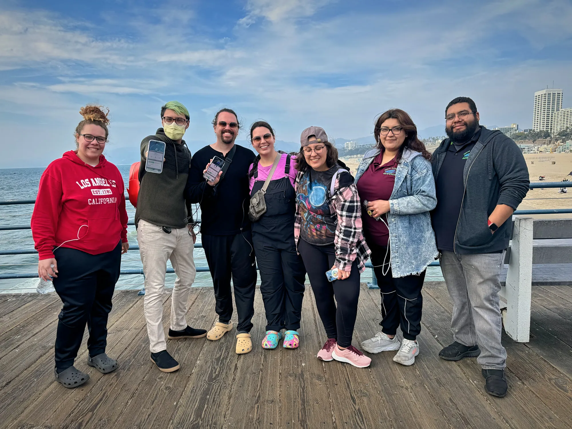 Seven friends standing together on the wooden deck of the Santa Monica Pier, with the beach and the hotels along the cliffs behind them.