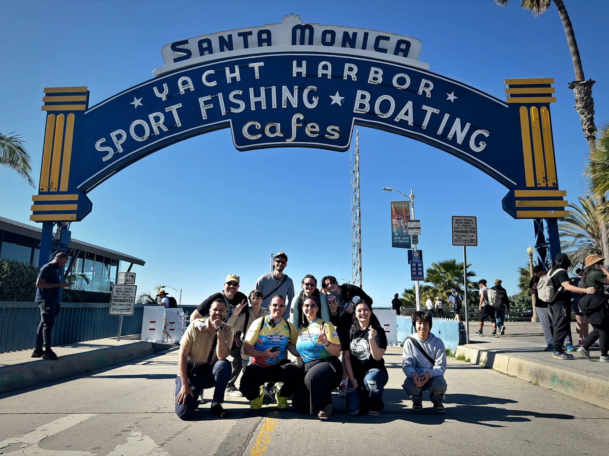 A big group of friends posing under the blue Santa Monica Yacht Harbor arch at the entrance to the pier on a sunny day.