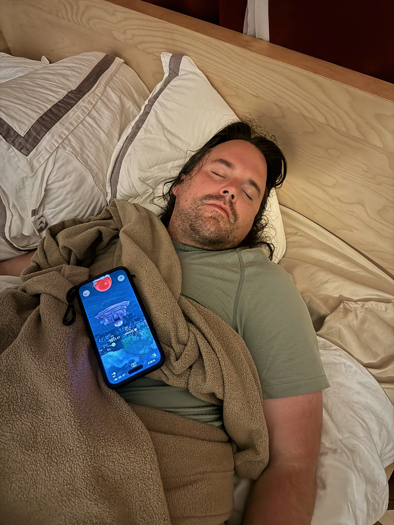 A man with long dark hair asleep in bed under a fleece blanket, with a phone still lit up lying on top of the blanket.