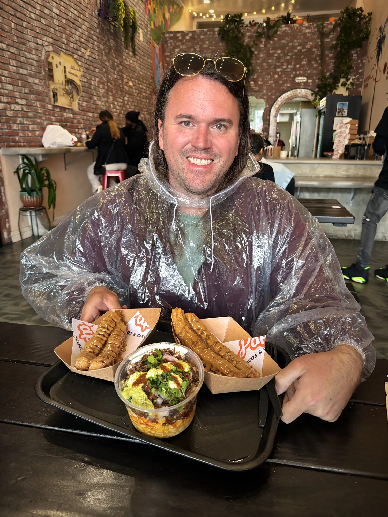 A smiling man in a clear plastic poncho holding a tray of churros and a taco bowl at a restaurant table in Santa Monica.