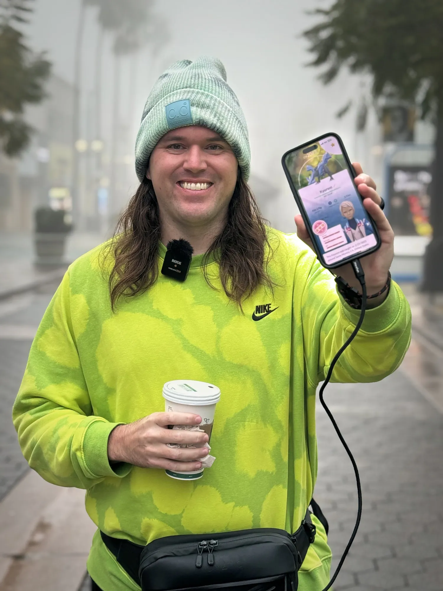 A man in a neon green sweatshirt and mint beanie holds up a coffee and a phone showing a Kyurem caught during Pokémon GO Tour weekend, on a foggy morning in Santa Monica.