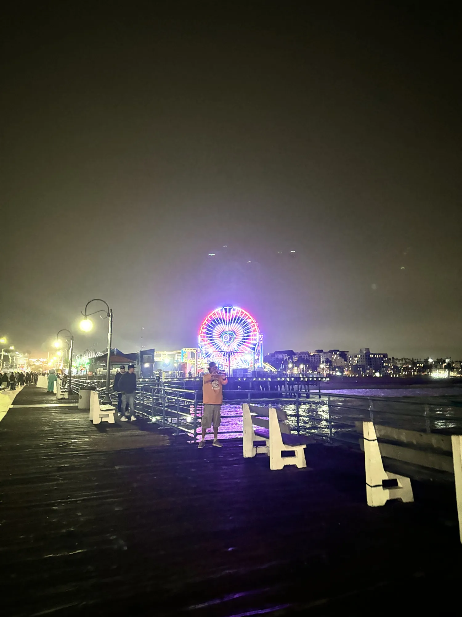 The lit-up Pacific Wheel at the end of Santa Monica Pier at night, seen down the wooden boardwalk with white benches and street lamps.