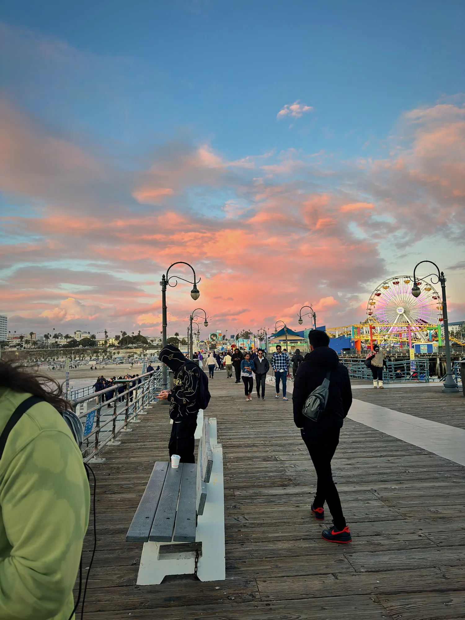 The wooden boardwalk of the Santa Monica Pier under a pink sunset sky, with the Pacific Park Ferris wheel at the far end.