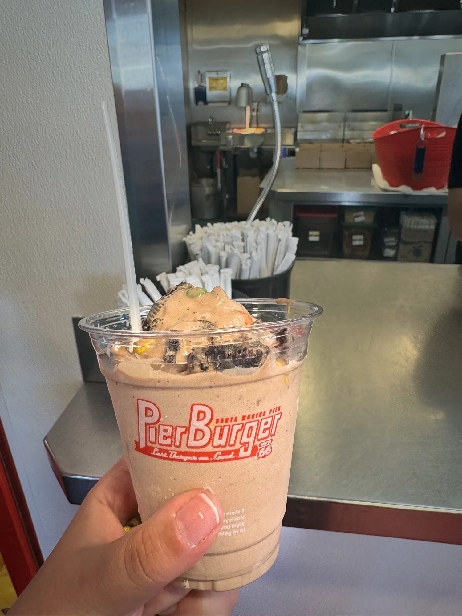 A hand holding a chocolate milkshake topped with whipped cream and cookie pieces in a Pier Burger cup at the counter.