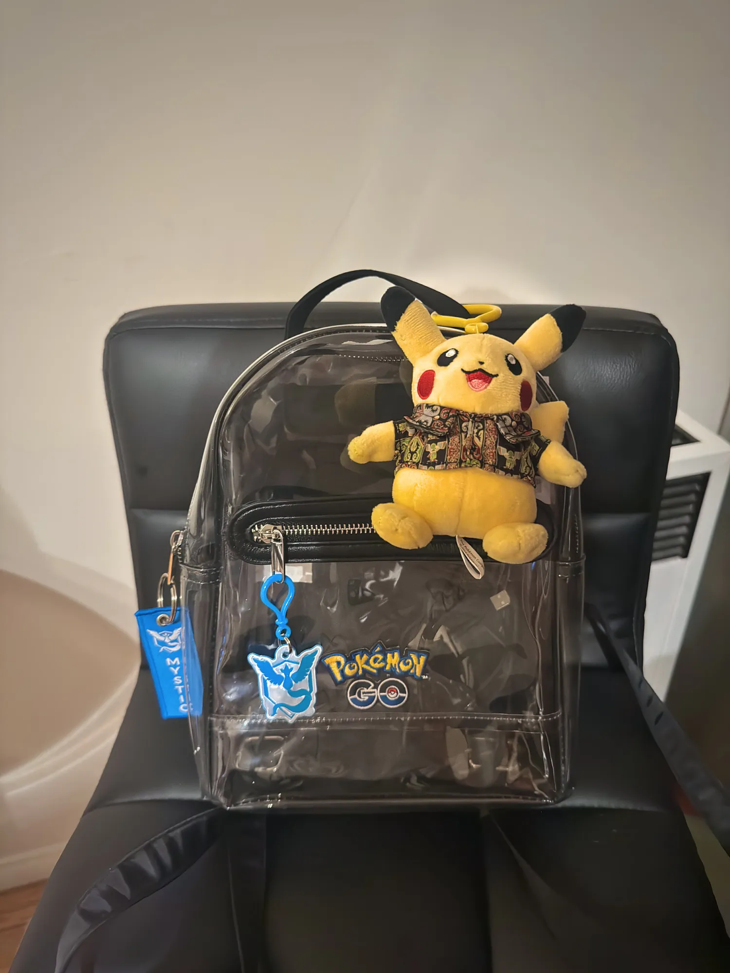 A small clear backpack with a Pikachu plush clipped to the top and a Team Mystic Pokémon GO keychain on the side, sitting on a black chair.