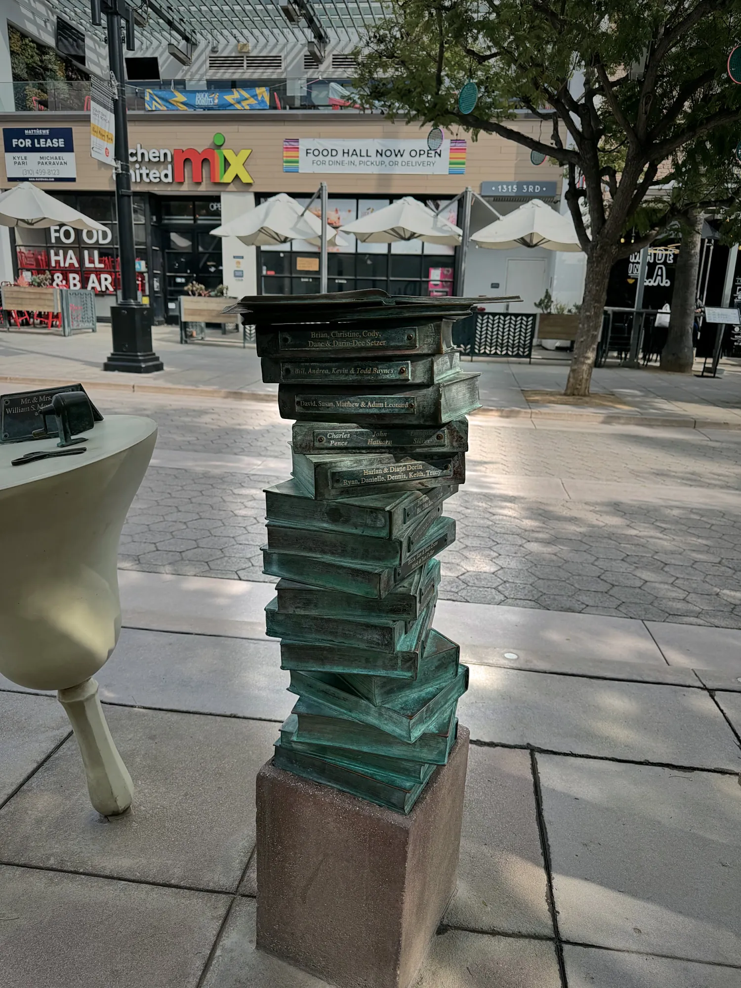 A bronze sculpture of a tall stack of books on the Third Street Promenade in Santa Monica, with shops behind it.