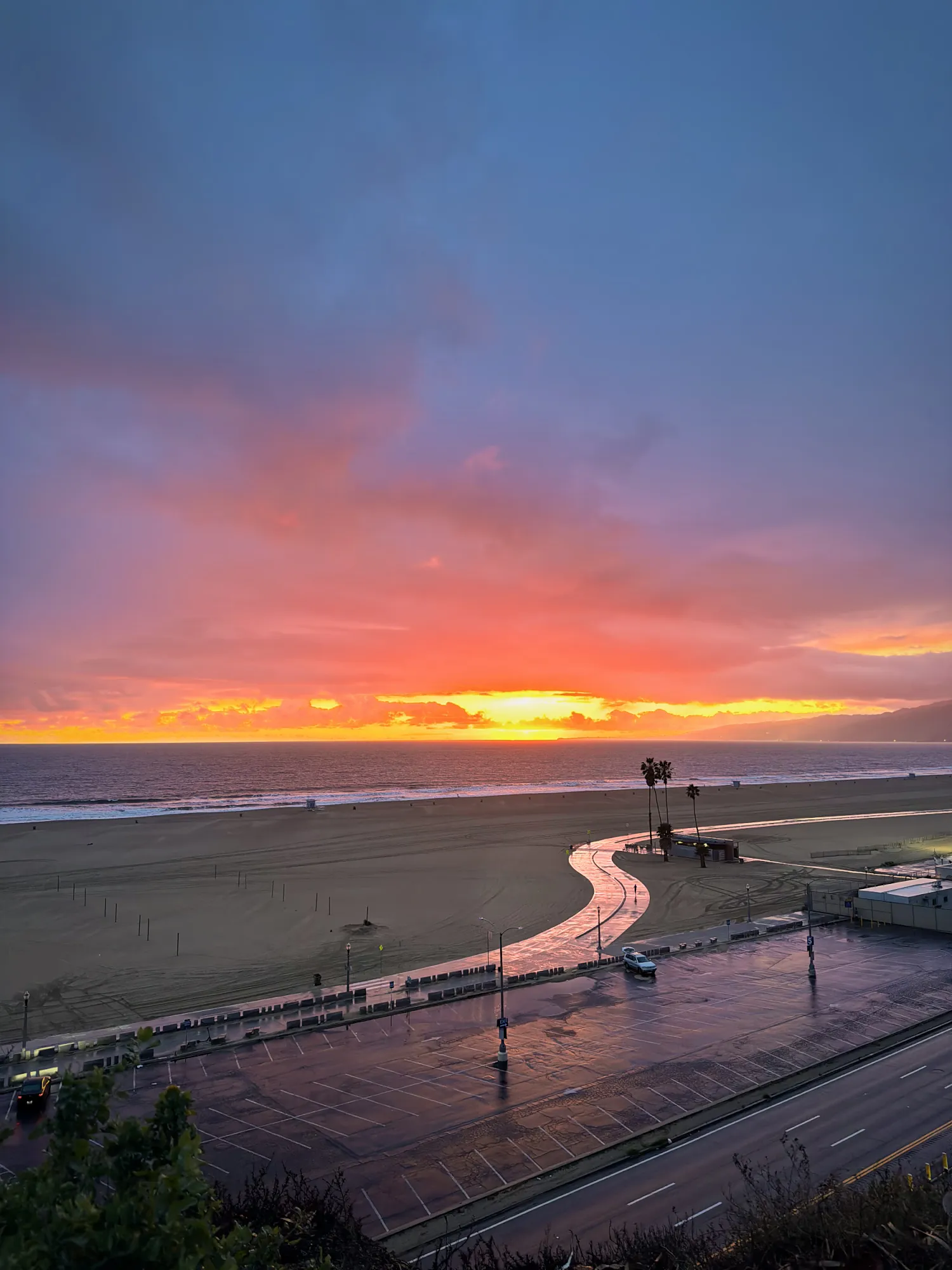 An orange and pink sunset over the ocean and the empty beach in Santa Monica, with the wet beach path curving across the sand below the cliffs.