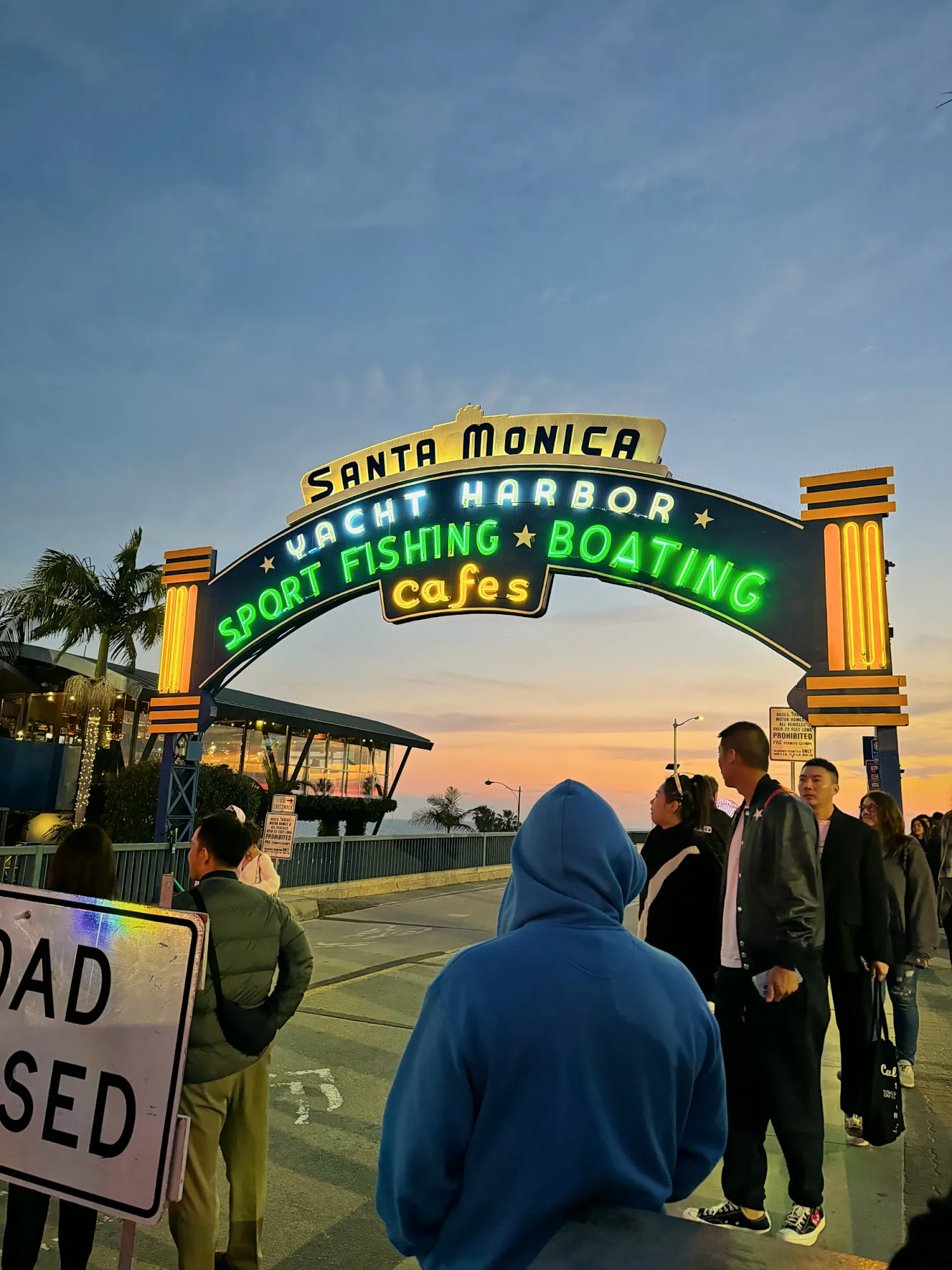 A crowd gathered under the neon-lit Santa Monica Yacht Harbor arch at the entrance to the pier at dusk.