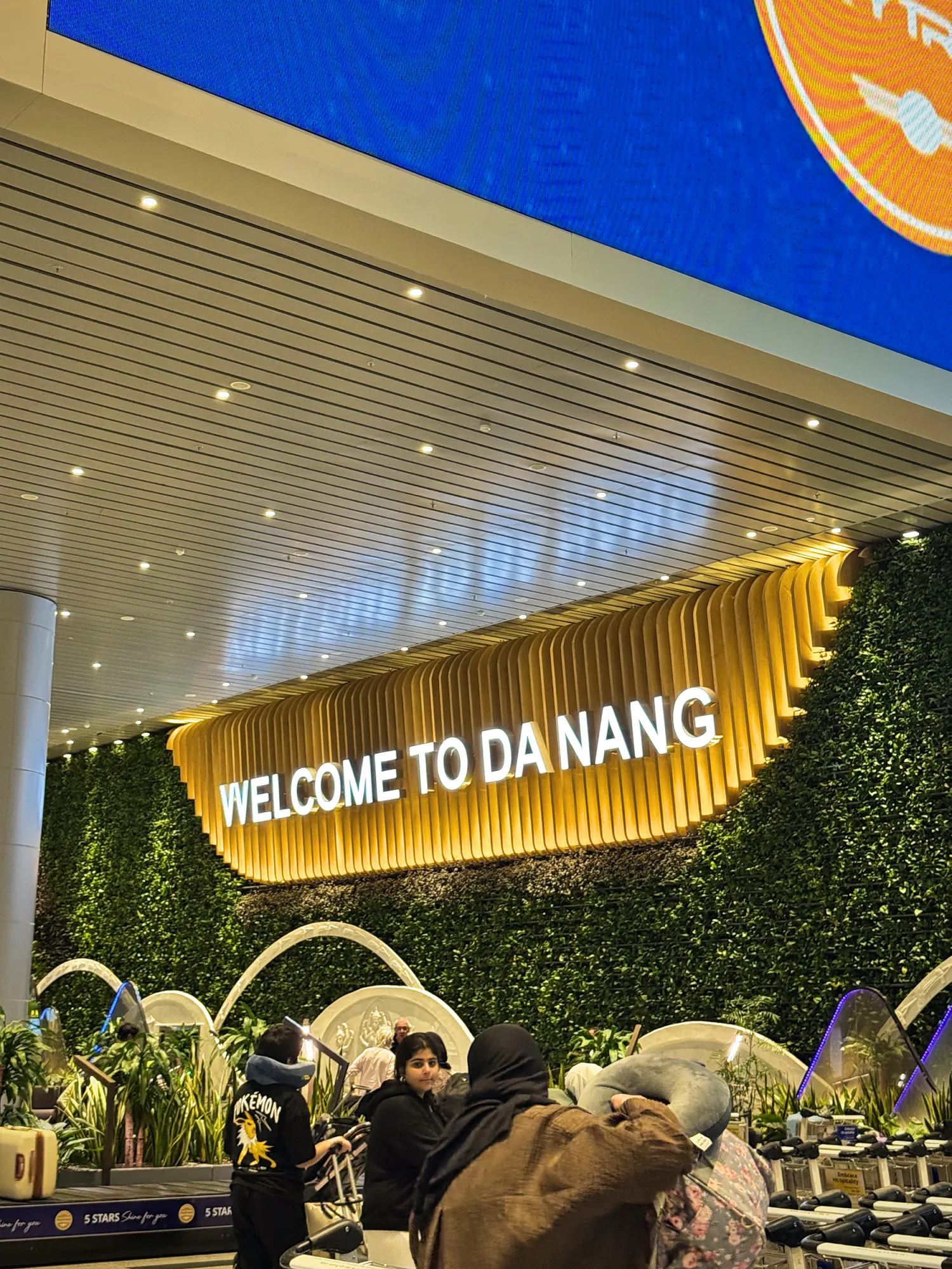 Welcome to Da Nang sign lit up above a green wall inside Da Nang International Airport, with travelers walking below.